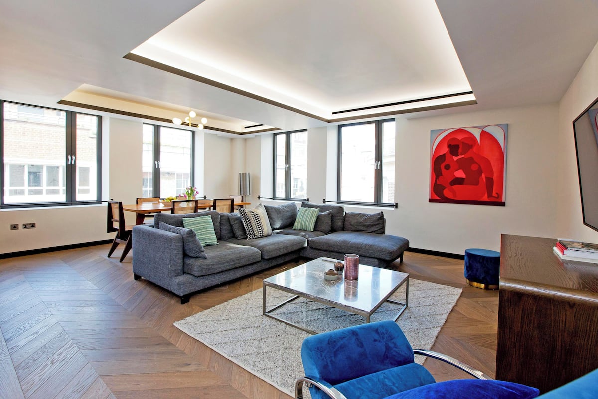 Ein modernes Wohnzimmer mit einem Ecksofa, einem Glastisch und einem blauen Sessel. Es gibt einen Essbereich mit Tisch und Stühlen, und ein großes Fenster sorgt für natürliches Licht.