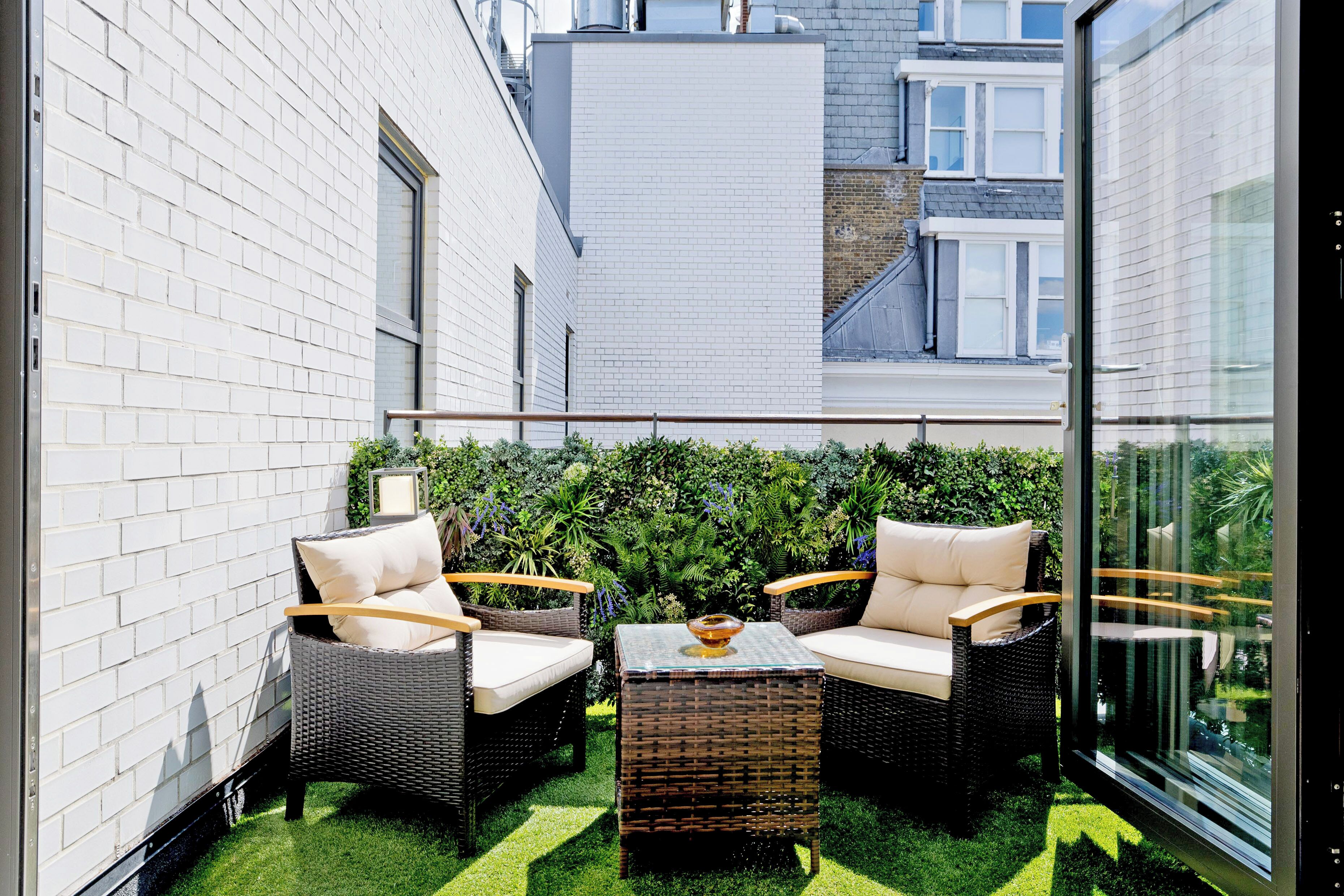 A balcony with wicker furniture, a glass table, and a green hedge wall.
