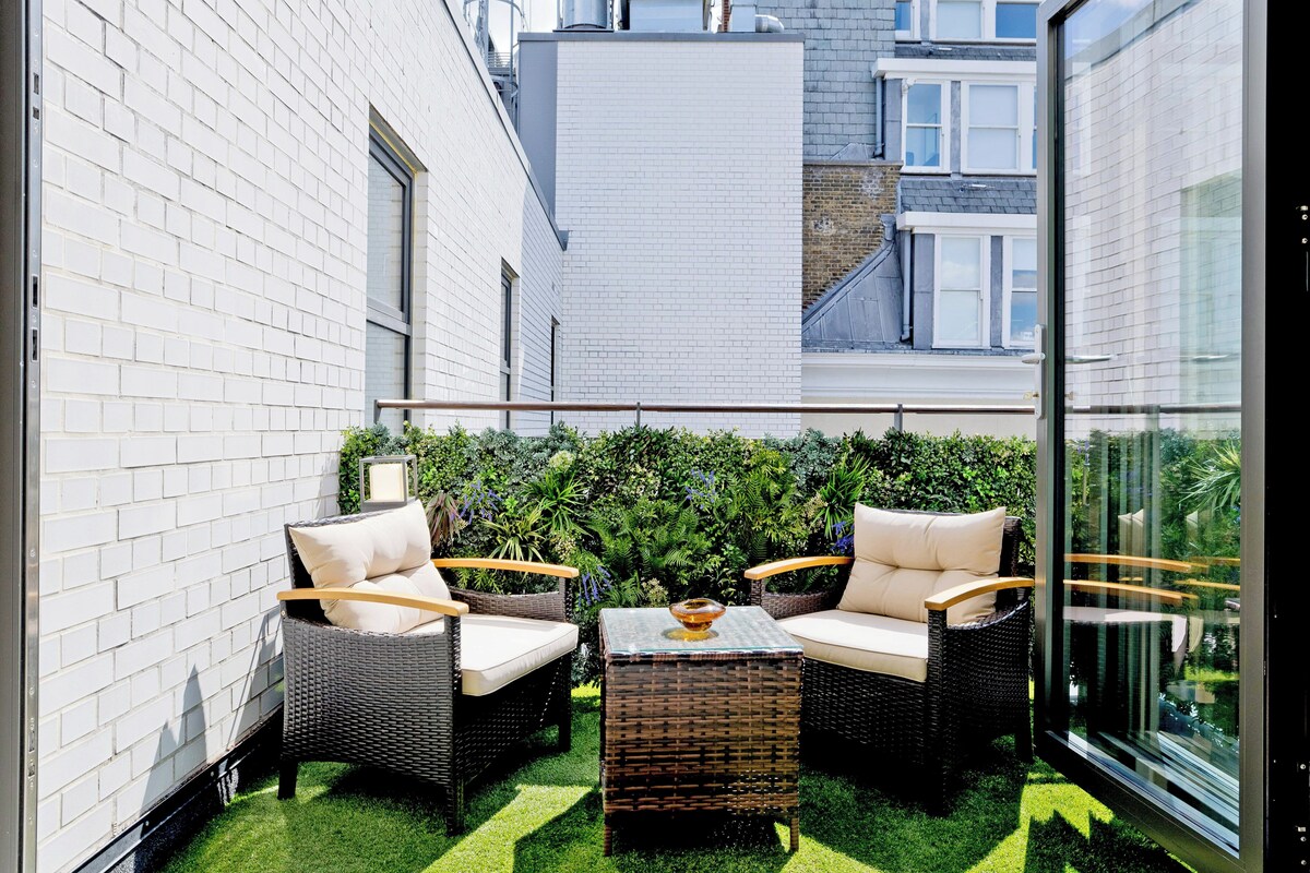 A balcony with wicker furniture, a glass table, and a green hedge wall.