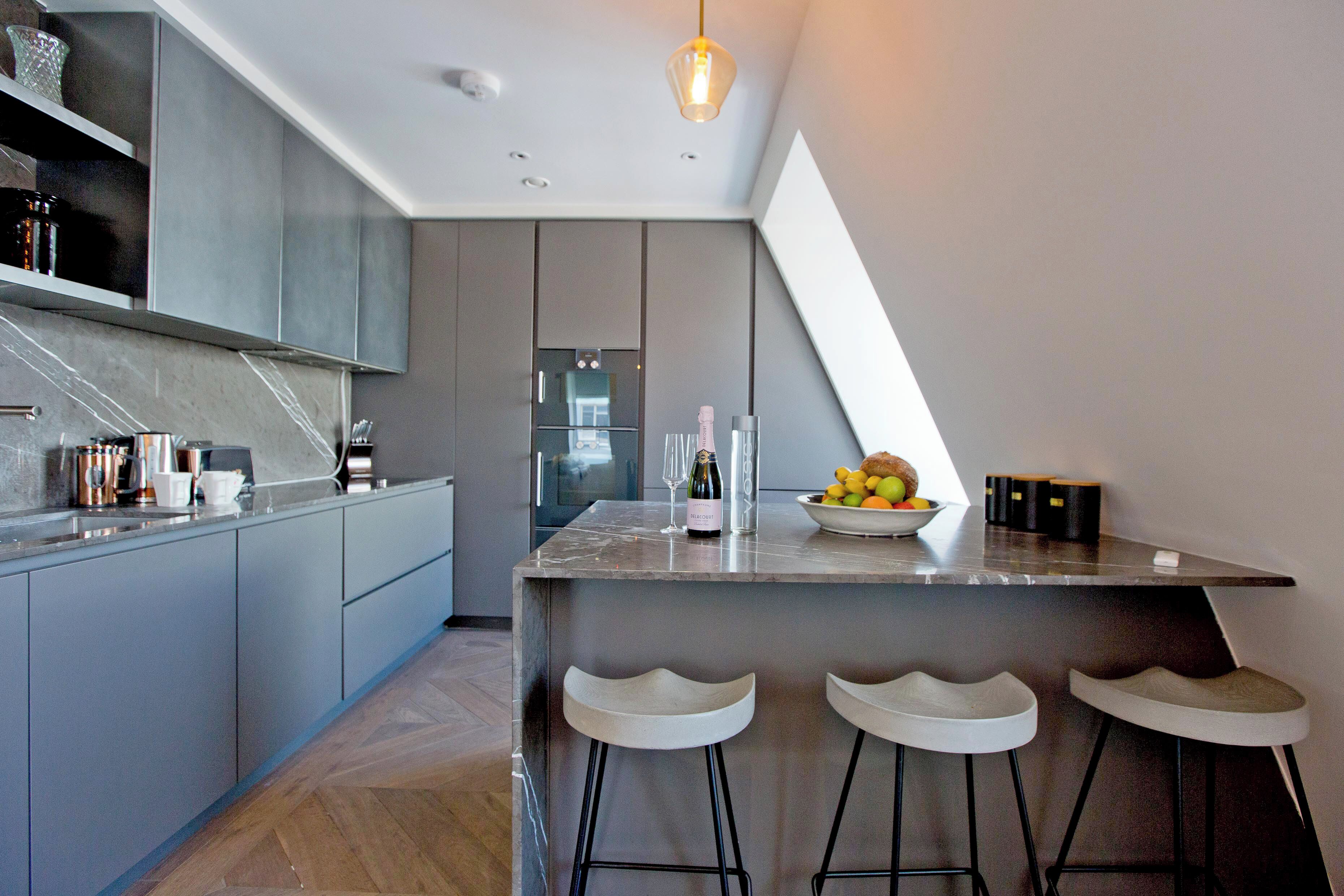 A modern kitchen with grey cabinets, a marble countertop, and a built-in oven. There are three bar stools and a bowl of fruit on the counter.