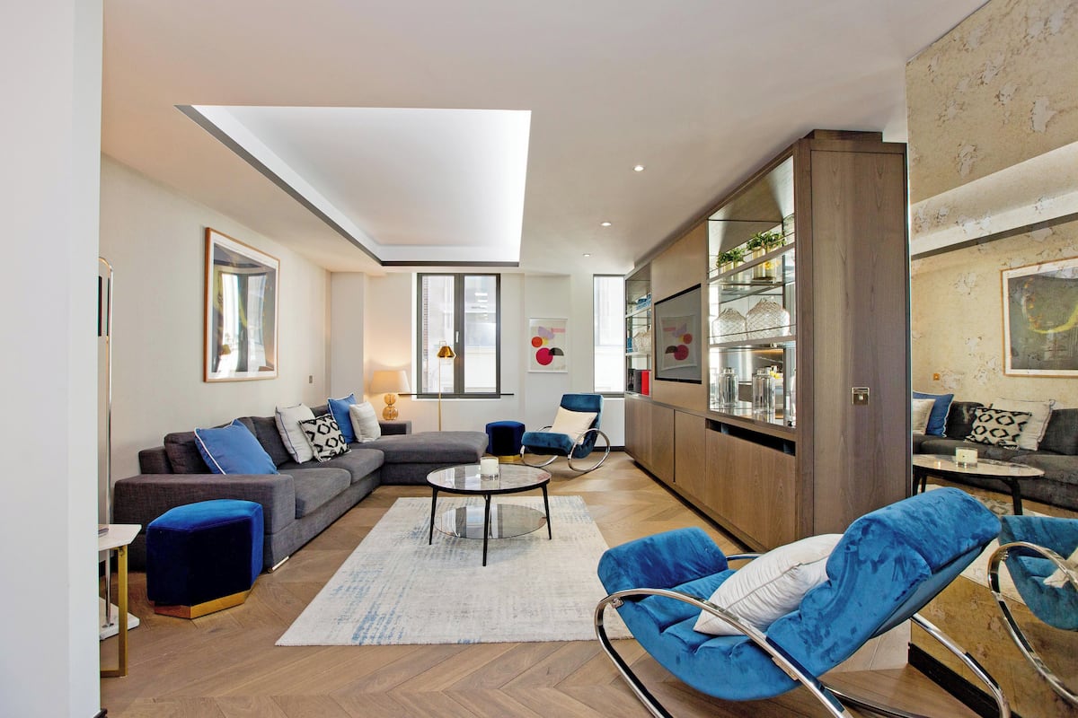 Ein modernes Wohnzimmer mit einer Couch, einem Hocker und blauen Sesseln, ein Essbereich mit Tisch und Stühlen sowie eine Küche mit Kühlschrank und Regalen.