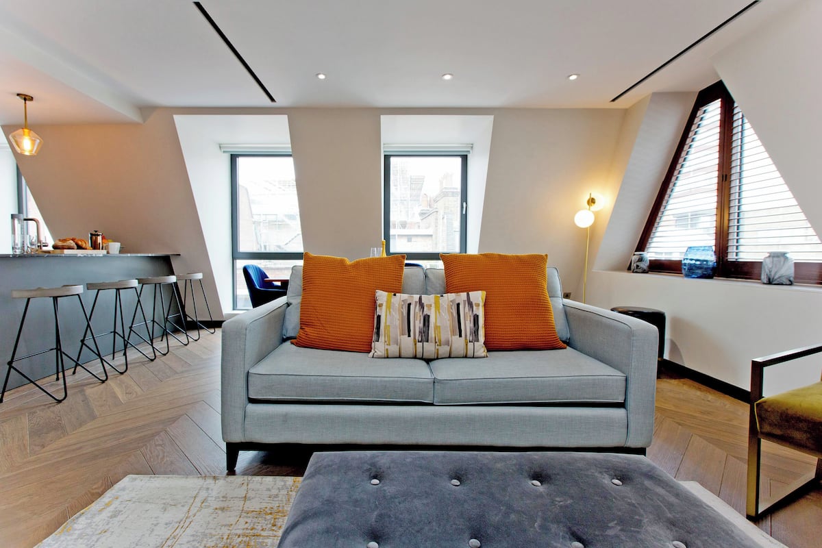 Ein modernes Wohnzimmer mit einem grauen Sofa, orangefarbenen Kissen und einem grauen Hocker. Zwei Fenster mit Jalousien, eine Küchenzeile mit Barhocker und eine Pendelleuchte sind ebenfalls vorhanden.