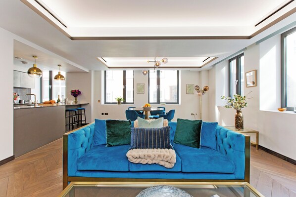 A modern living room with a blue sofa, a glass coffee table, and a dining area in the background.