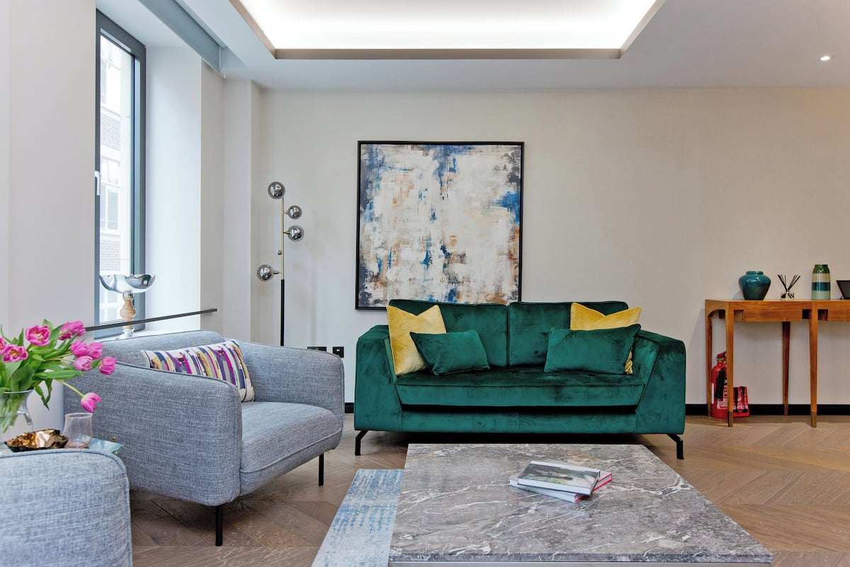 Ein modernes Wohnzimmer mit einem grauen Sessel, einem grünen Sofa mit gelben Kissen, einem Marmortisch und einem großen, abstrakten Gemälde an der Wand.