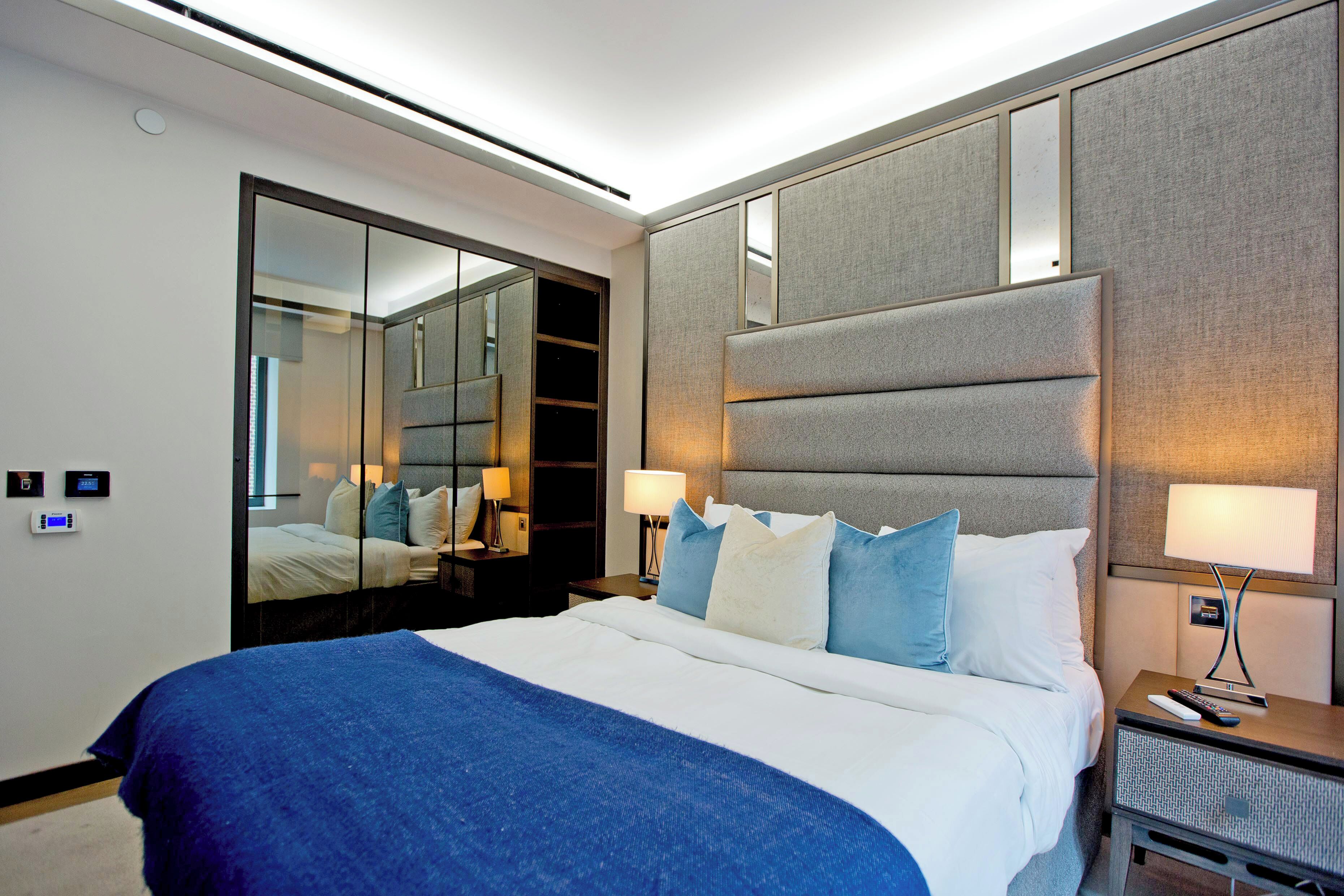 A modern hotel room with a large bed, a mirror, a nightstand, and a wall-mounted panel.