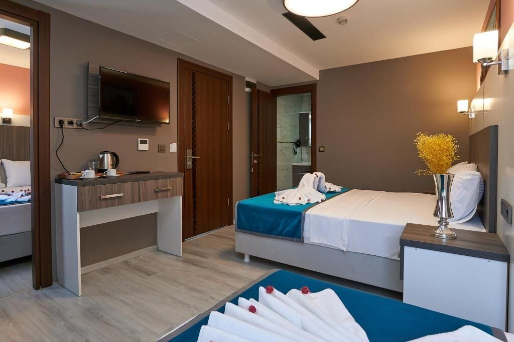 Quarto de hotel com cama, mesa de cabeceira, escrivaninha com cadeira, TV fixada na parede e banheiro visível através de uma porta aberta.