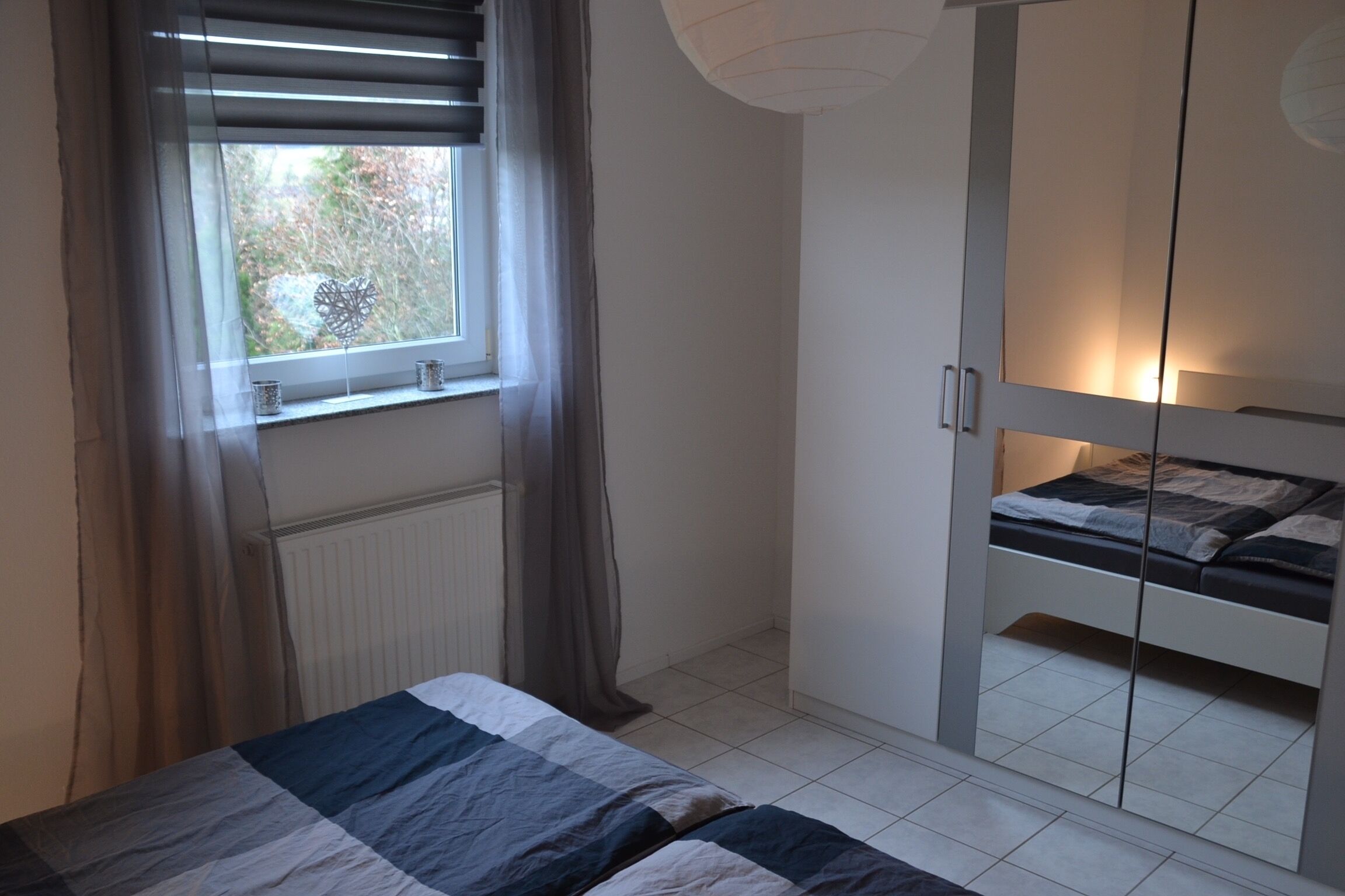 A bedroom with a bed, a window with curtains, a radiator, and a wardrobe with a mirror.
