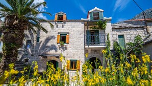 A stone building with multiple windows and balconies, surrounded by yellow flowers and a palm tree. - Holiday house Pivcevi (Podgora)