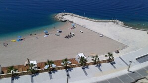 An aerial view of a beach with clear blue water, a sandy area with umbrellas and lounge chairs, and a wooden deck with palm trees. - Holiday house Pivcevi (Podgora)