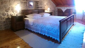 A bedroom with a stone wall, wooden ceiling beams, a bed with a white lace bedspread, a nightstand, a lamp, and a framed picture on the wall. - Holiday house Pivcevi (Podgora)