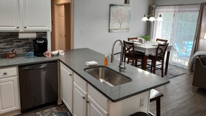 A modern kitchen with a built-in dishwasher, a coffee maker, and a sink. There is a dining area with a table and chairs, and a large window with curtains. - Luxury! 2 king beds and 2 full bathrooms (Manteno)
