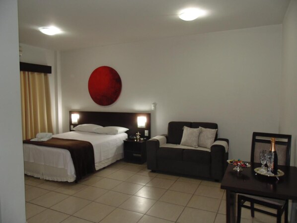 A hotel room with a bed, a sofa, a table with a bottle and glasses, and a red circular decoration on the wall.