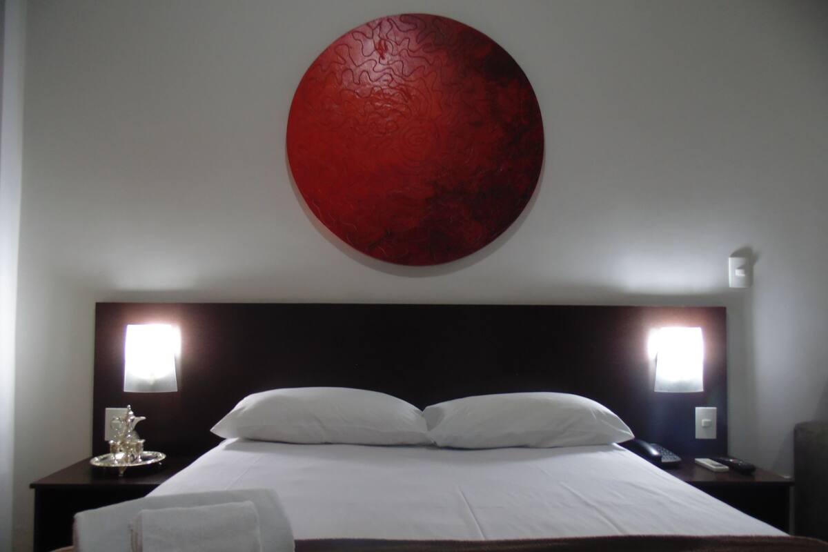 A hotel room with a large bed, two bedside tables, a red wall decoration, and a window with blinds.
