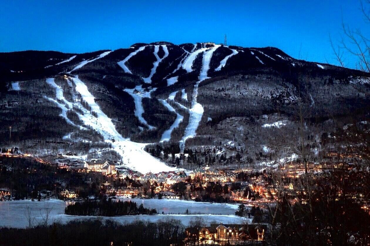 Une montagne au pied de laquelle se trouve une station de ski, avec une ville aux bâtiments illuminés et un ciel nocturne dégagé.