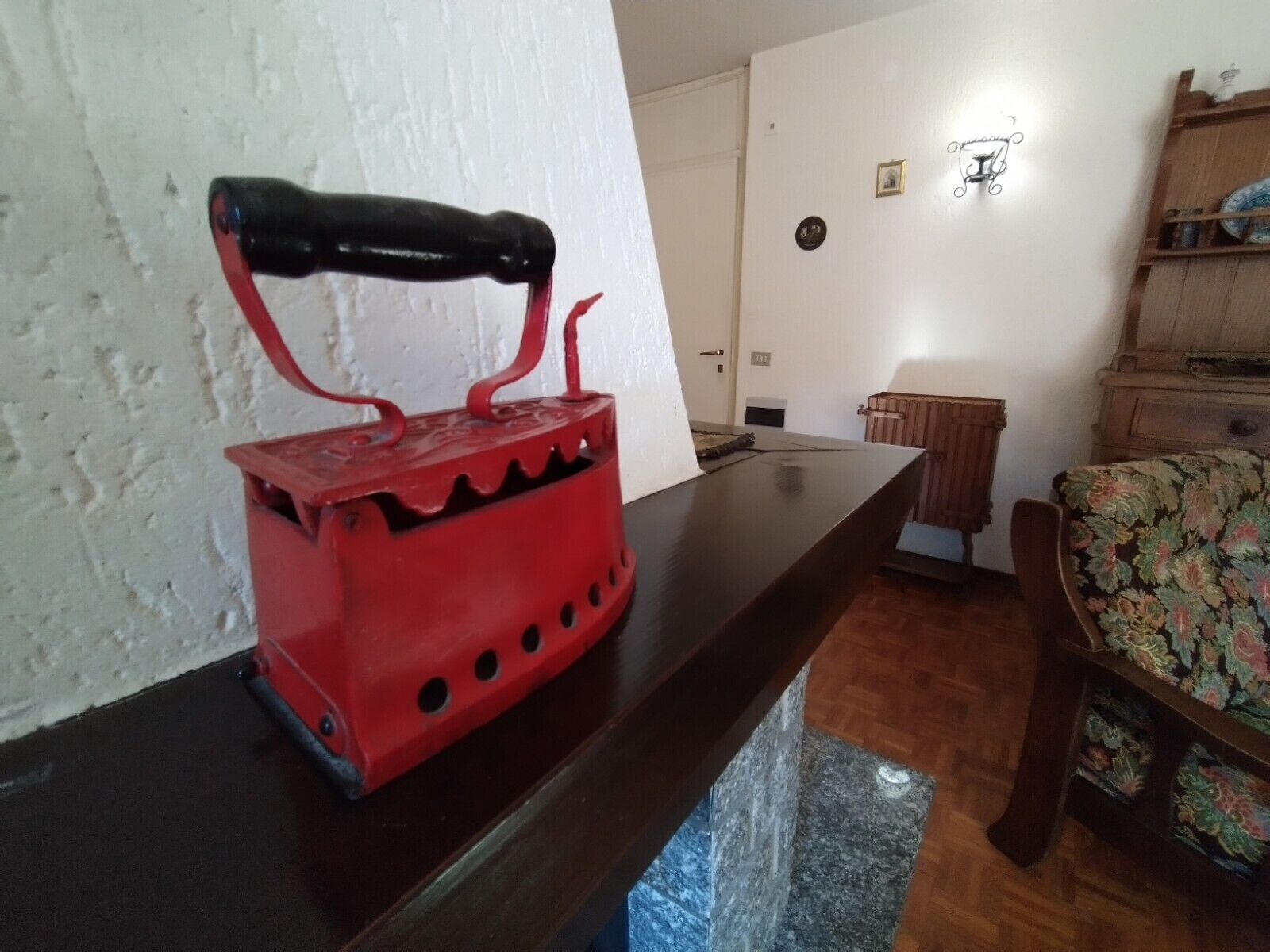 A red kettle on a wooden table in a room with a white wall, a wooden cabinet, and a floral chair.