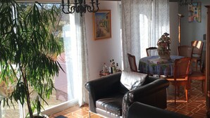 A living room with a leather sofa, a dining table with chairs, a chandelier, and a large window with curtains.