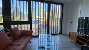 A modern living room with a glass-top coffee table, a brown leather sofa, a TV on a wooden stand, and large windows with a city view. - New luxury 3bedroom townhouse cityview (South Melbourne)