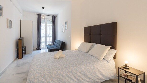 Un dormitorio con una cama grande, una mesita de noche con lámpara, una silla y una ventana con cortinas.
