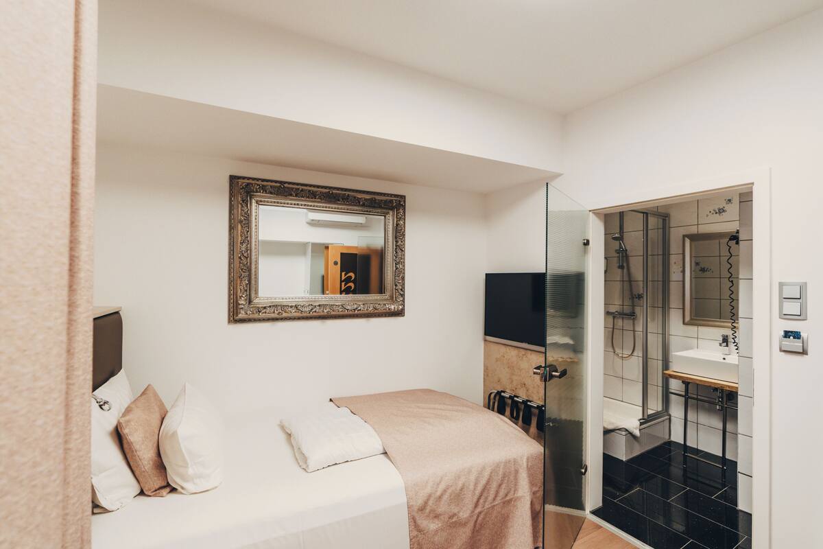 A hotel room with a bed, a mirror, a television, a shower, and a sink.
