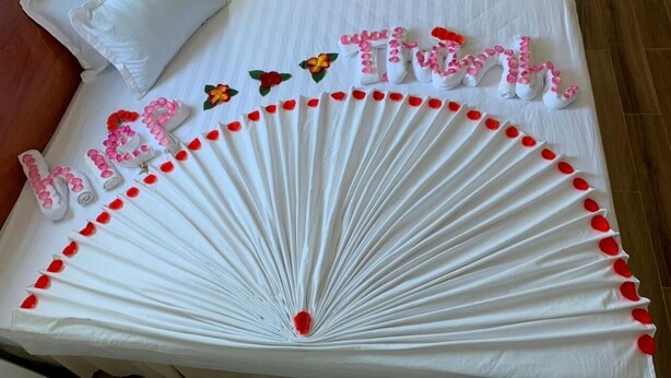Una cama con edredón blanco, una almohada y elementos decorativos, incluyendo un arreglo de tela blanca con detalles rojos y las palabras 'happy birthday' en letras rosadas.