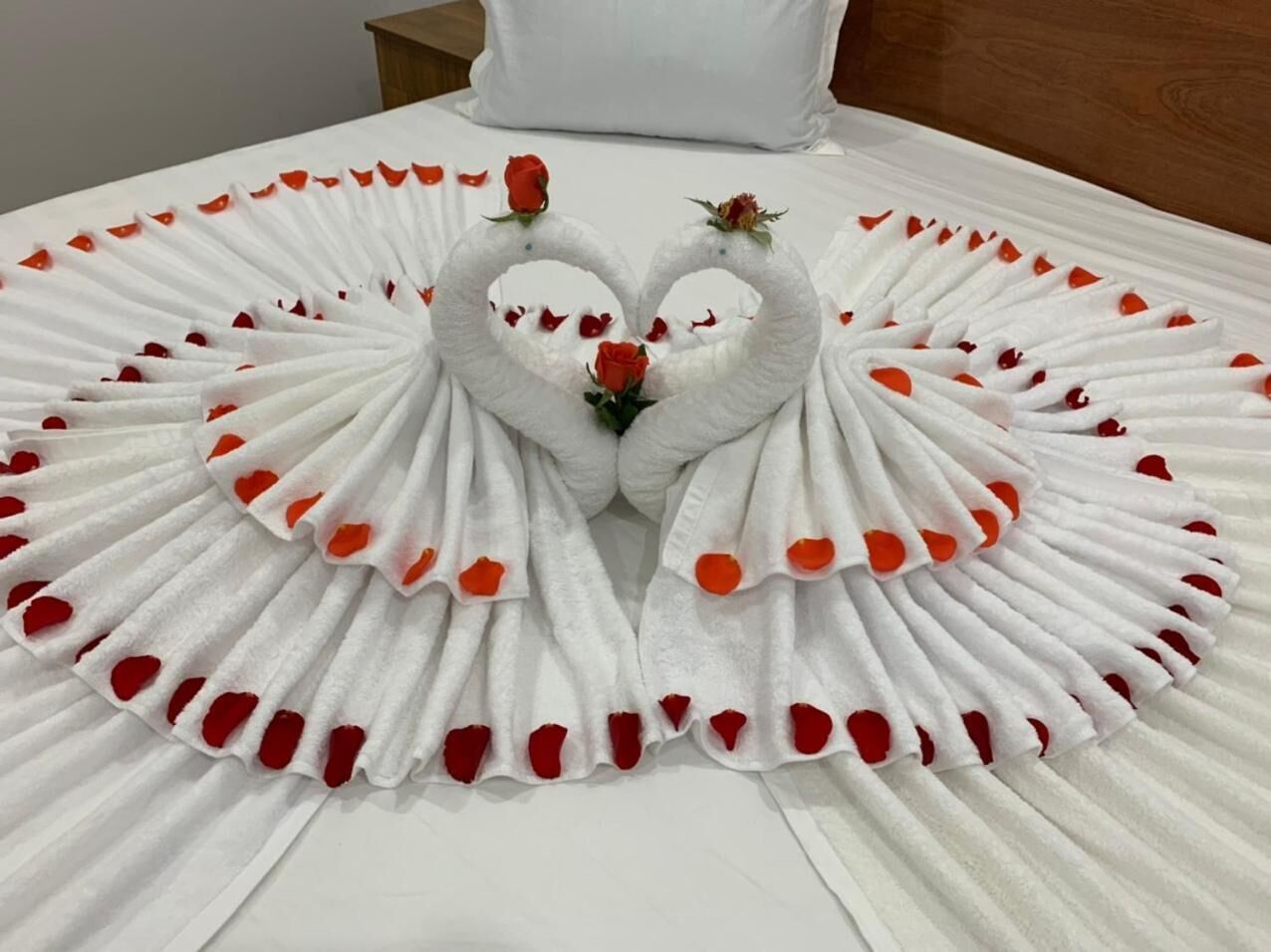 Una cama con sábana blanca y una composición decorativa de toallas blancas y pétalos de rosa roja formando un corazón en el centro.