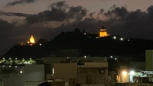 Vista nocturna de una colina con una estructura iluminada, posiblemente un monumento o templo, y edificios circundantes con luces.