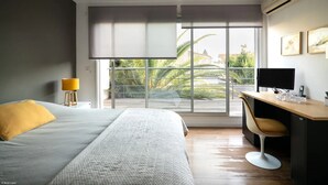 Ein modernes Hotelzimmer mit großem Fenster, einem Bett, einem Schreibtisch mit Stuhl und einem kleinen Tisch mit Lampe.