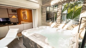 Ein moderner Außenbereich mit Whirlpool, einer Couch, Lounge-Stühlen, einem Fernseher und einem großen Fenster mit Blick auf Grünflächen.