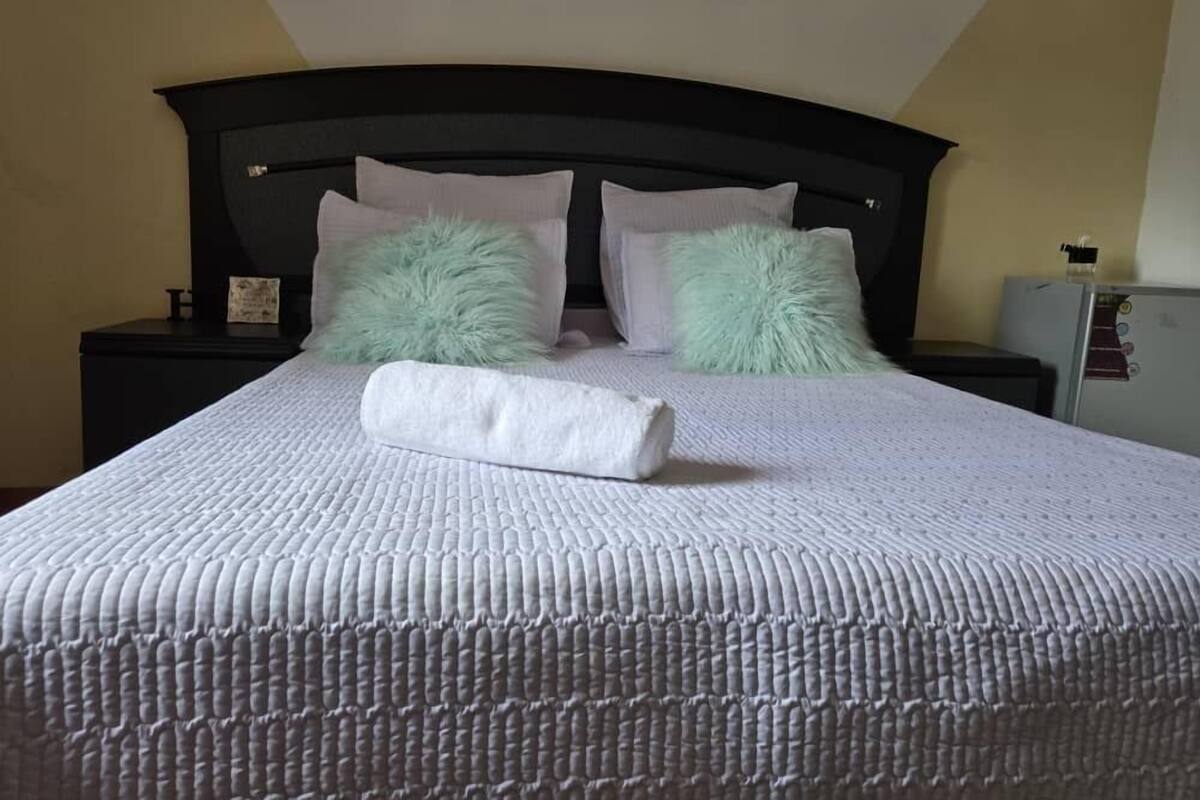 A neatly made bed with a white quilt and two fluffy pillows, a dark headboard, and a small nightstand with a lamp.