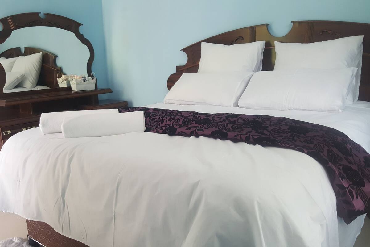 A neatly made bed with white linens and a purple floral blanket, a wooden headboard, and a small bedside table with a mirror and a basket.