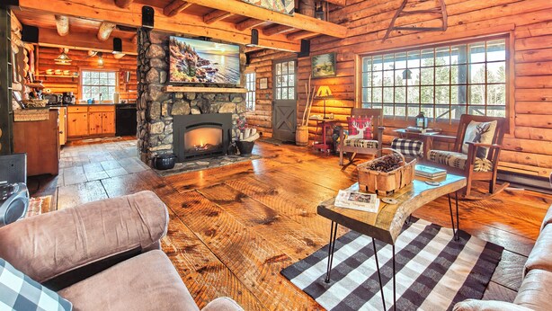A cozy log cabin interior with a stone fireplace, wooden floors, a dining area, and large windows. - 40 Acre Forested Privacy w/ Deeded Beach Access Near Acadia Schoodic (Prospect Harbor)