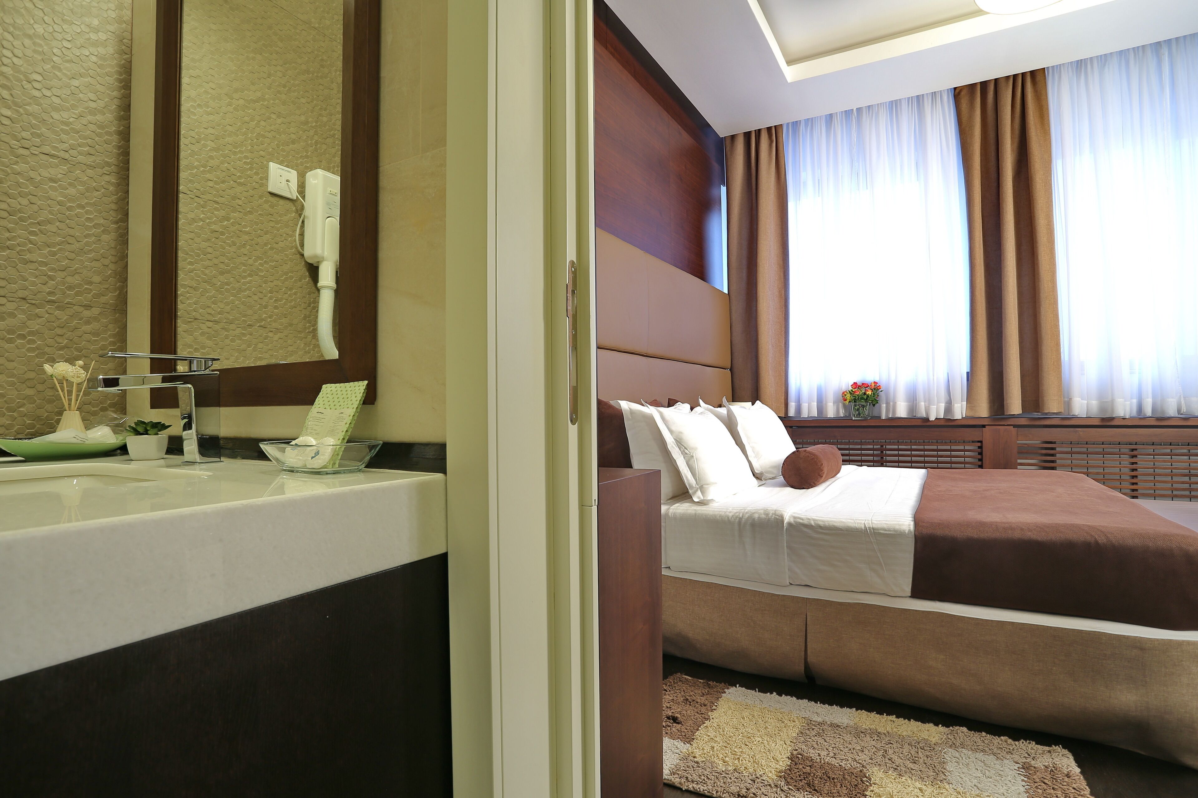 A hotel room with a large bed, a bathroom with a mirror, and a window with curtains.