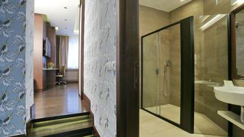 A modern hotel bathroom with a glass-enclosed shower, a white sink, and a wooden staircase leading to a room with a TV and a flower vase.