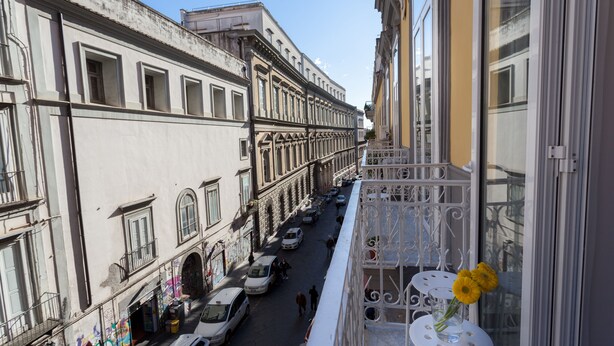 A narrow street with buildings on either side, a balcony with a white railing, and a vase with flowers. - Manuela Apartments Piazza San Domenico in the historic center (Napoli)