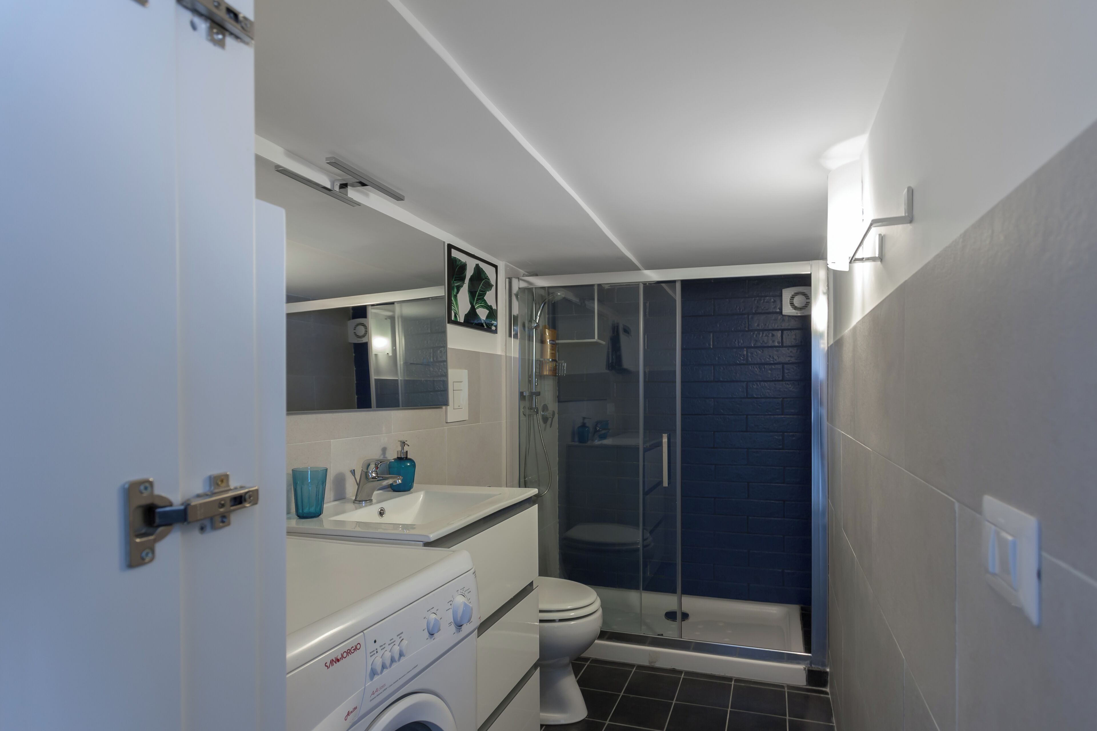A compact bathroom with a white sink, a toilet, a glass shower enclosure, and a washing machine.