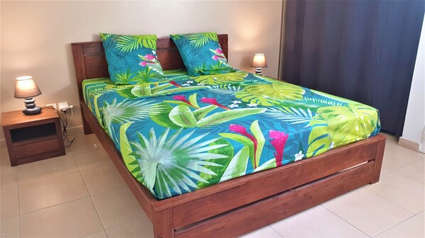 A bed with a tropical floral bedspread, two matching pillows, a wooden headboard, a nightstand with a lamp, and a power outlet.