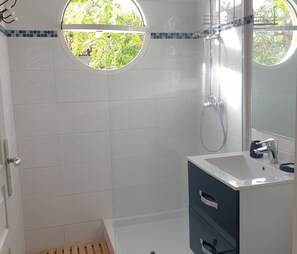 Ein Badezimmer mit weißen Fliesen, einer Dusche, einem Waschbecken mit Spiegel und einem Fenster mit Blick ins Grüne.