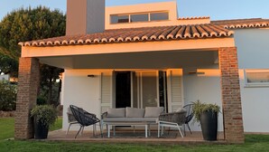 Ein modernes Haus mit überdachter Terrasse, zwei Stühlen, einer Couch und einem großen Topfpflanzen.