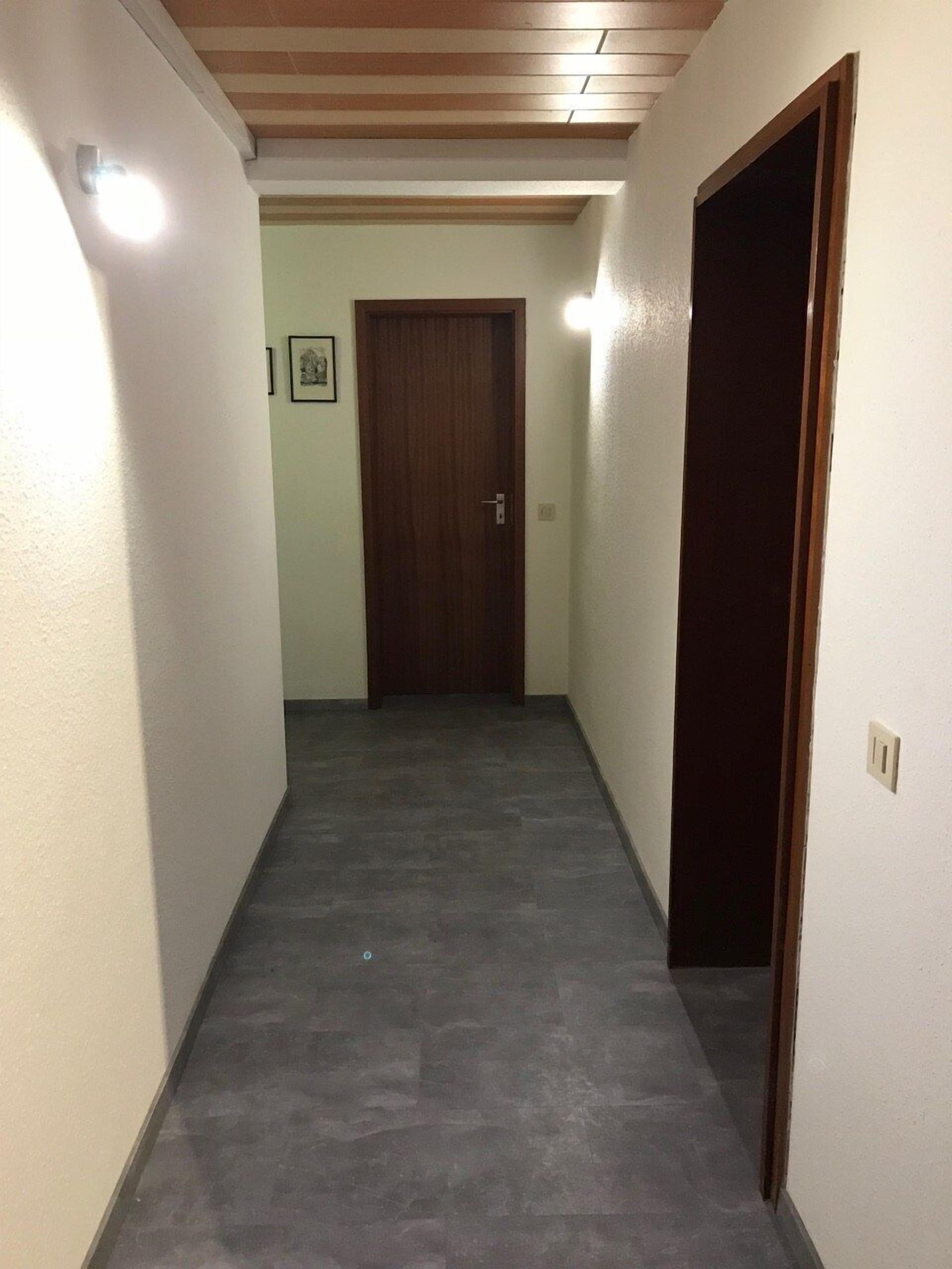 A narrow hallway with a wooden ceiling and a dark floor. A wooden door is visible at the end of the hallway.