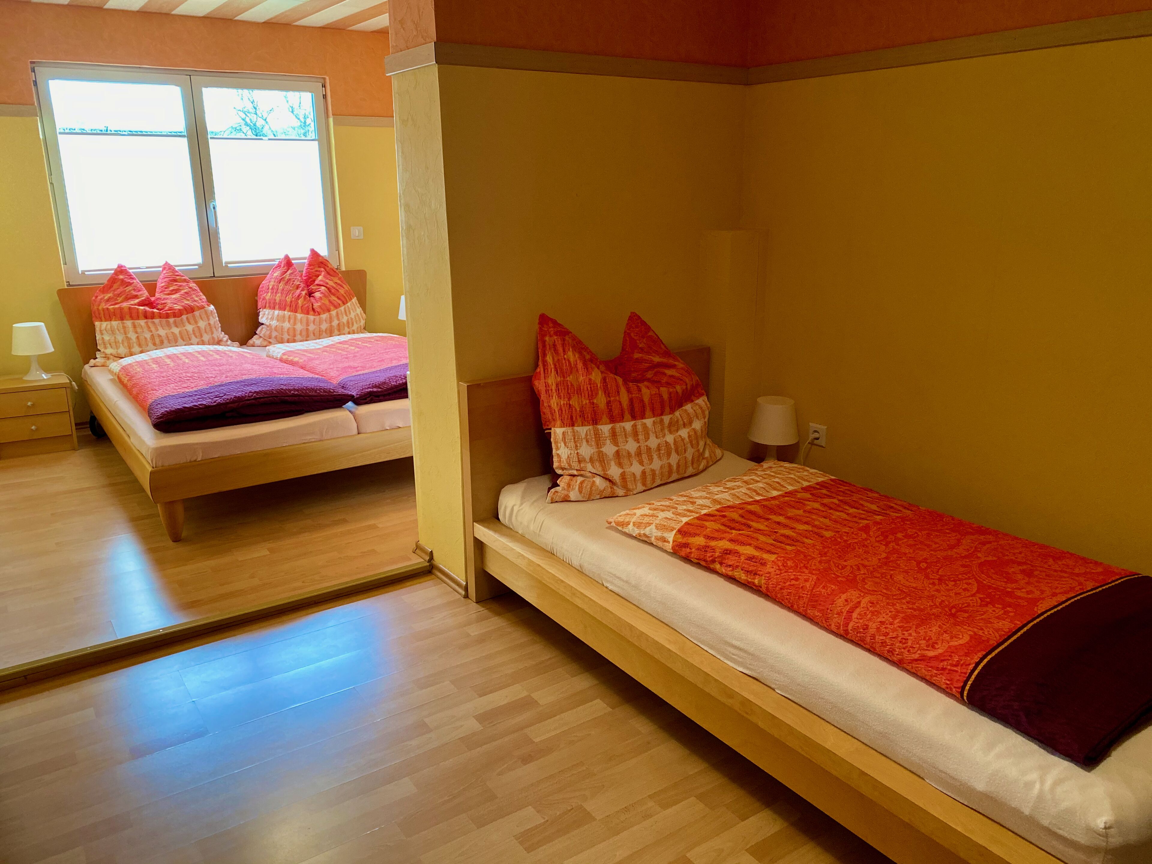 Une chambre avec deux lits, chacun agrémenté de draps et d’oreillers à motifs. Une fenêtre laisse entrer la lumière naturelle, et la chambre est dotée d’un plancher en bois.