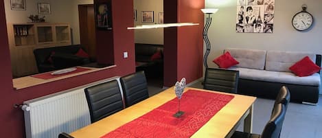 Un comedor con una larga mesa, sillas negras y un mantel rojo. Se ve un sofá con cojines rojos al fondo y una pared decorada con cuadros.