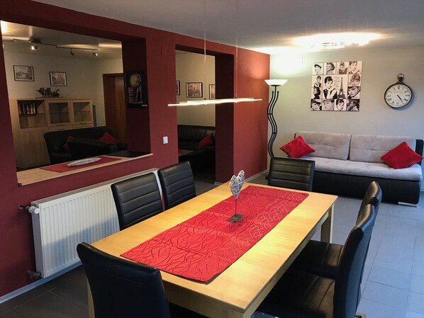 Un coin repas avec une longue table, des chaises noires et un chemin de table rouge. On aperçoit un canapé avec des coussins rouges à l’arrière-plan, ainsi qu’un mur orné de tableaux encadrés.