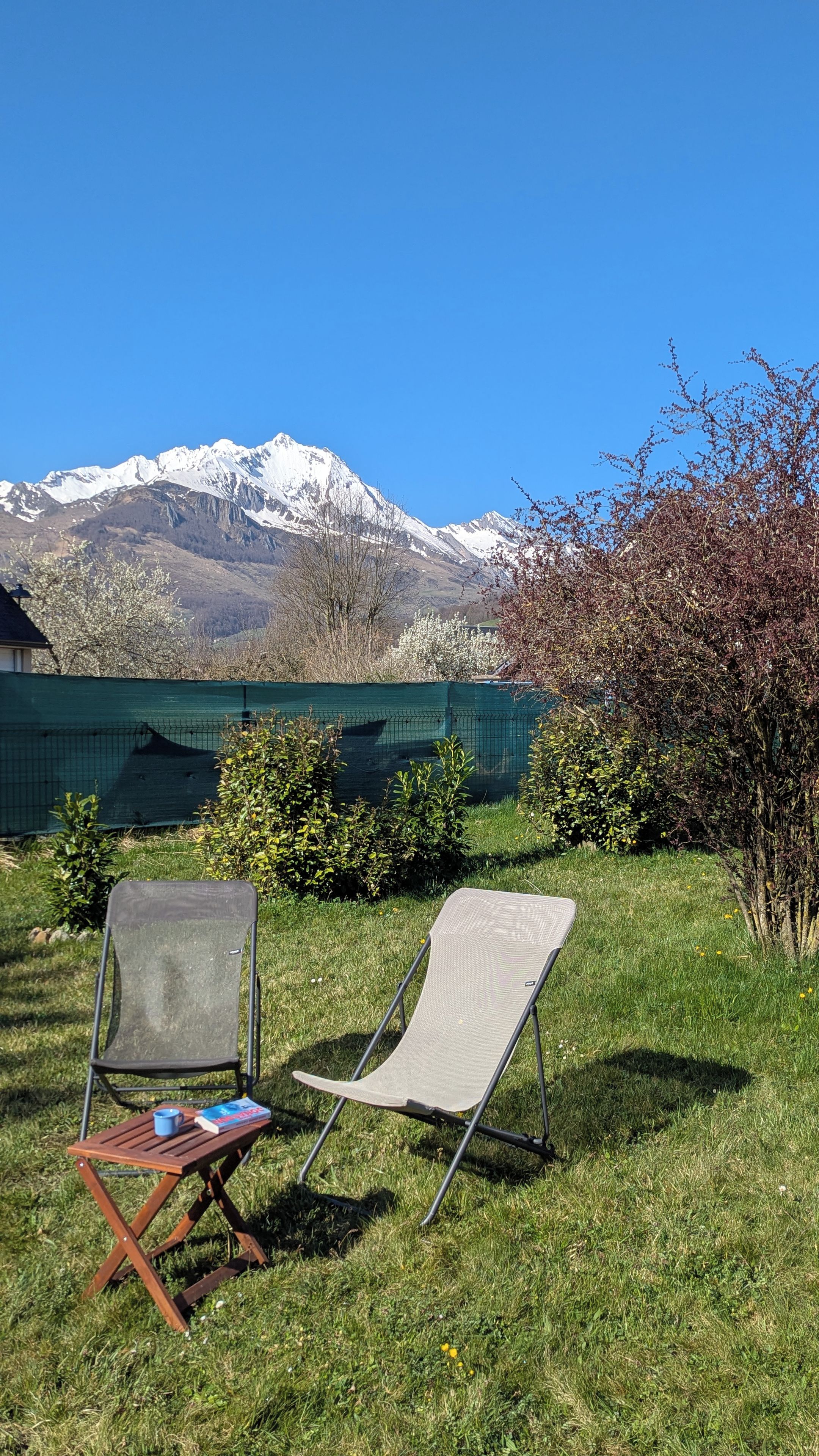 Two outdoor chairs with a book and a mug, a green fence, and a mountain range in the background.