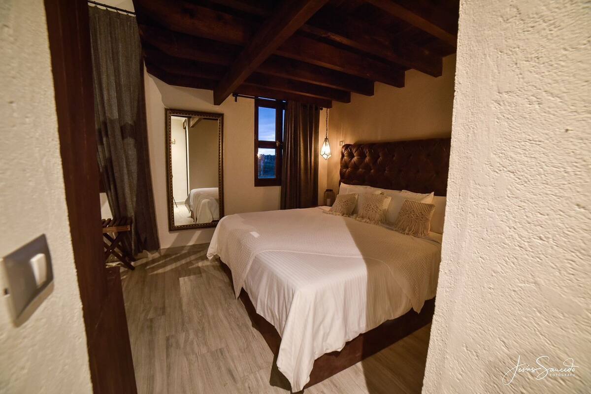 A bedroom with a large bed, wooden beams, a mirror, and a window with curtains.