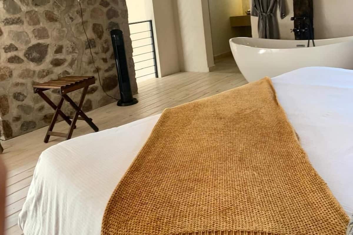 A bedroom with a large bed, a television mounted on a stone wall, a small stool, and a bathtub in the background.