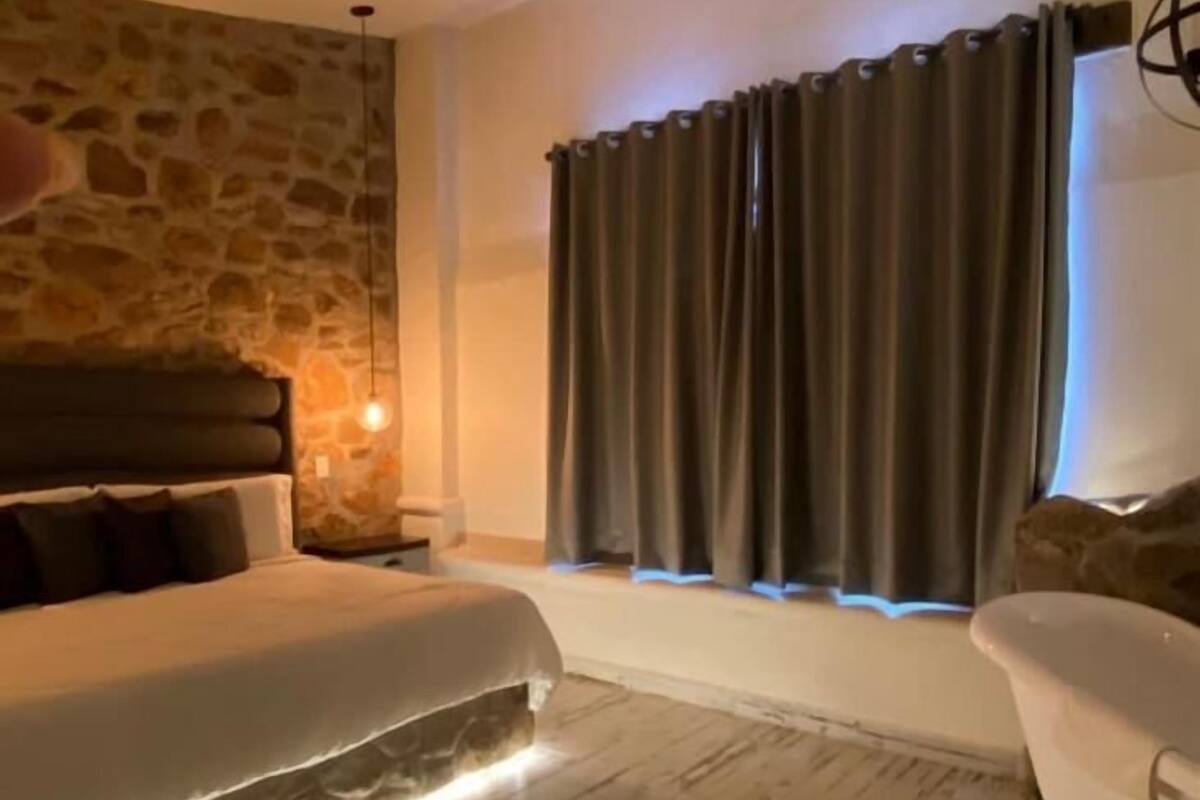 A bedroom with a stone wall, a bed with a headboard, a bedside table, a nightstand, a window with curtains, and a white chair.