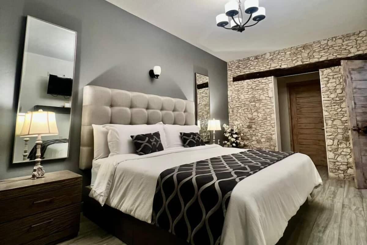 A bedroom with a large bed, a nightstand with a lamp, a wall-mounted mirror, and a stone accent wall.