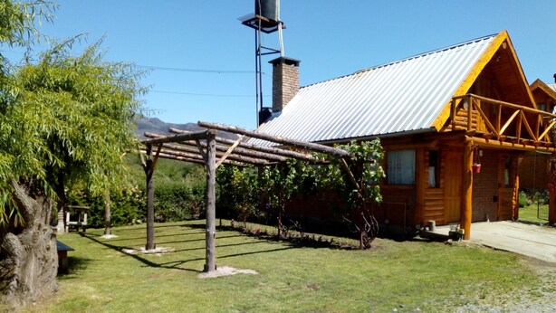 A rustic wooden cabin with a metal roof, a wooden pergola, and a gravel driveway.