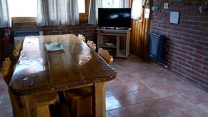 A wooden dining table with chairs, a television on a stand, and a brick wall with various decorations.
