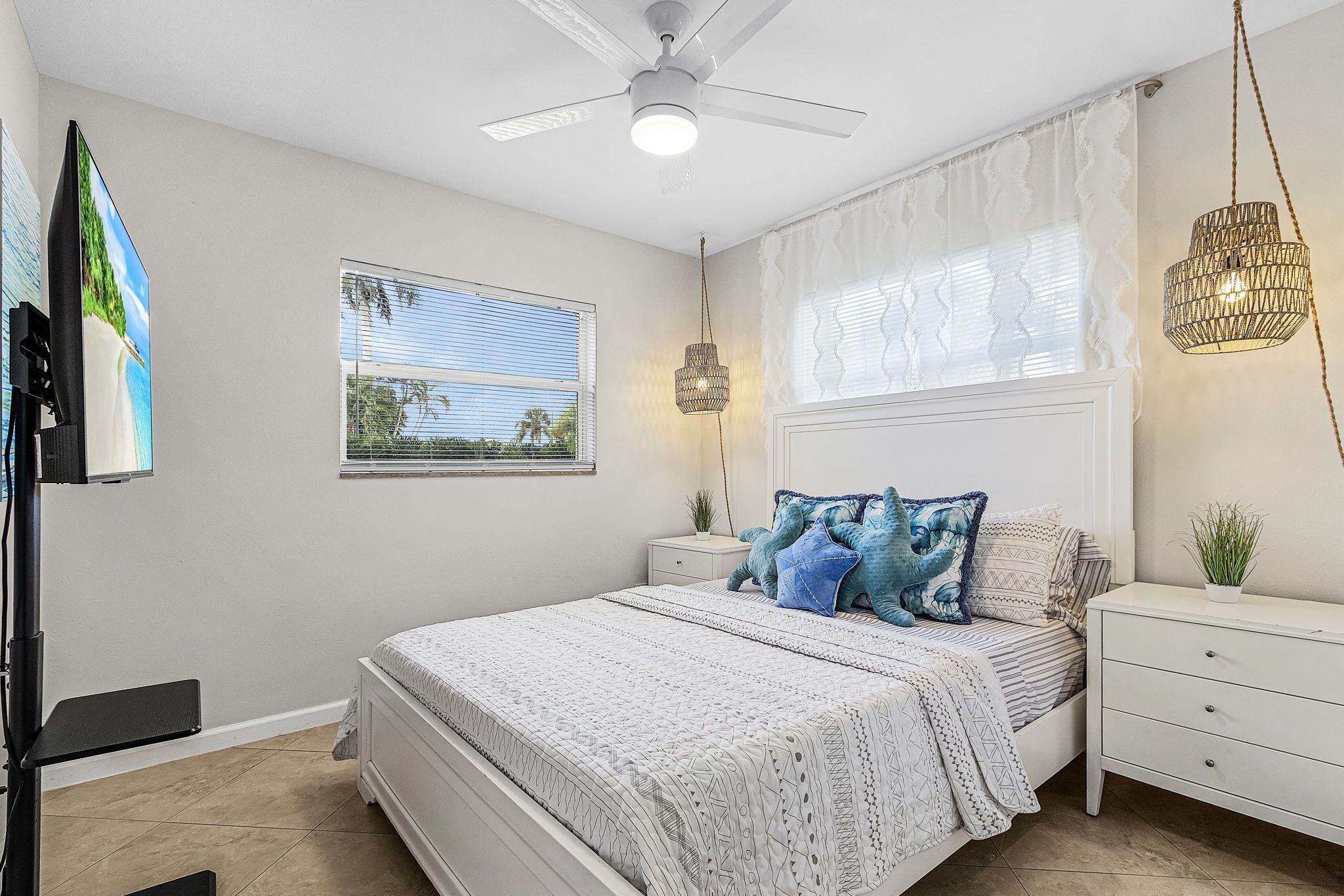 A bedroom with a bed, a ceiling fan, a window with blinds, a bedside table with a lamp, and a TV mounted on the wall.